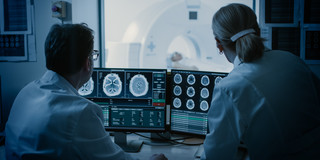 A man and a woman sit in the control room during an MRI and watch the images on a screen.