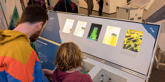 Im vorderen Teil des Bildes ist eine Station mit Fächern zu sehen. Die Rückseiten eines Kindes und eines Mannes, welche sich die Station anschauen, befinden sich vorne links im Bild. Im Hintergrund ist ein Ausstellungsraum und weitere Personen zu sehen.