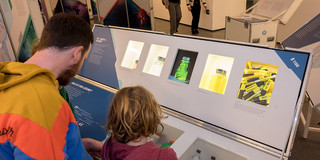 Im vorderen Teil des Bildes ist eine Station mit Fächern zu sehen. Die Rückseiten eines Kindes und eines Mannes, welche sich die Station anschauen, befinden sich vorne links im Bild. Im Hintergrund ist ein Ausstellungsraum und weitere Personen zu sehen.