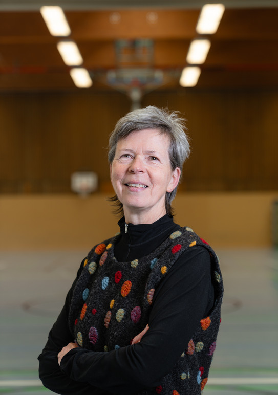 Portrait photo of Anke Hellwig: The photo shows a woman with short gray hair standing in a gym with her arms crossed.
