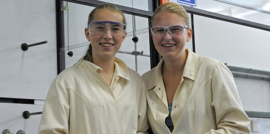 2 Schülerinnen mit Schutzbrillen und in weißen Kitteln stehen in einem Labor