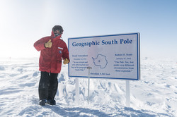 Geografischer Südpol Ein Mann in dicker roter Jacke neben einer Tafel im Schnee, auf der "Geographic South Pole" steht