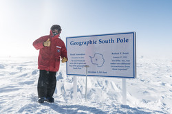 A man in a thick red jacket standing next to a plaque in the snow that reads "Geographic South Pole"