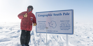 Ein Mann in dicker roter Jacke neben einer Tafel im Schnee, auf der "Geographic South Pole" steht