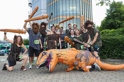 Mehrere als Neandertaler verkleidete Personen halten eine Keule in der Hand. Im Vordergrund liegt eine als Dinosaurier verkleidete Person.