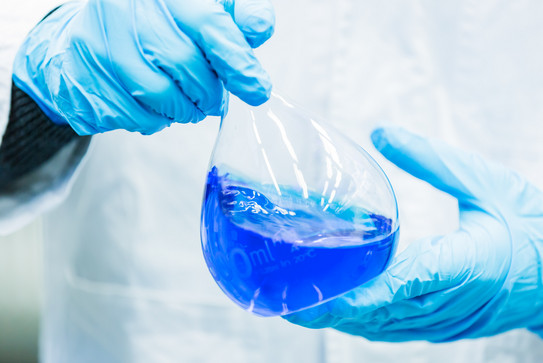 Detailaufnahme Messkolben Zwei Hände in blauen Gummihandschuhen halten einen Messkolben der mit blauer Flüssigkeit gefüllt ist.