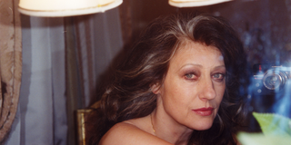 A portrait that an older lady with long black and gray hair has taken of herself in a mirror with a compact camera. She is looking through the mirror into the camera, which she is holding in her hand next to her head. Curtains and two bright lampshades can be seen around her in the room.