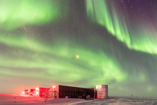 Amundsen-Scott South Pole Forschungsstation Ein zweistöckiges Gebäude mit vier Seitenflügeln im Schnee unter einem Nachthimmel mit grün leuchtenden Polarlichtern.