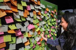 Foto: Eine Frau hängt ein Post-It an eine grüne Stellwand, an der viele bunte Post-Its kleben.