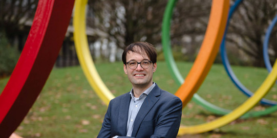 Ein Porträtbild von einem Mann, Prof. Lukas Buchheim, draußen vor bunten großen Stahlringen.