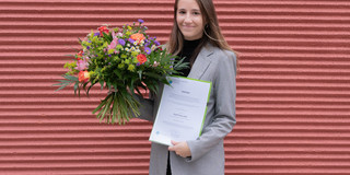 María Ángeles García Flores Eine junge Frau steht im Blazer vor einer Backsteinwand und hält einen Blumenstrauß und eine Urkunde in der Hand.