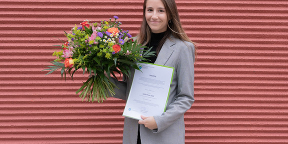 Eine junge Frau steht im Blazer vor einer Backsteinwand und hält einen Blumenstrauß und eine Urkunde in der Hand.