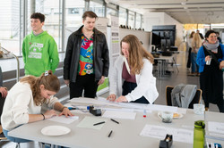 An einem Tisch im Rudolf-Chaudoire-Pavillon basteln zwei Studentinnen und ein Student an etwas aus weißen Papierblättern. Um sie herum stehen weitere Menschen, einer von ihnen hat einen grünen TU-Hoodie an.