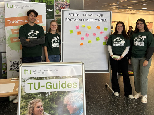 Vier Studierende der TU Dortmund stehen an einem Infostand mit dem Titel ‚Study Hacks für Erstakademiker*innen‘. Neben ihnen sind bunte Post-its auf einem Whiteboard und ein Aufsteller mit der Aufschrift ‚TU-Guides‘ zu sehen.