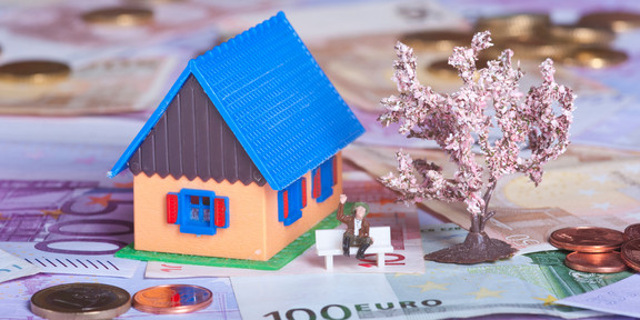 Zu sehen sind ein Miniaturhaus, ein Miniaturbaum und eine Miniaturbank, auf der eine Miniaturperson sitzt. Auf dem Boden liegen mehrere Geldscheine und Geldmünzen.