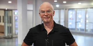 Portrait von Prof. Stefan Turek: Ein Mann mit schütterem, weißen Haar und Brille in einem schwarzen Poloshirt steht im Foyer des Audimax.