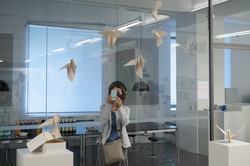 Ausstellung „Rundgang Kunst“ Eine Frau fotografiert mit einem Handy ein Kunstobjekt, dies besteht aus mehreren kleinen weißen Vögeln, die an der Wand herab hängen.