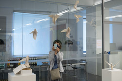 Eine Frau fotografiert mit einem Handy ein Kunstobjekt, dies besteht aus mehreren kleinen weißen Vögeln, die an der Wand herab hängen.