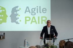 Das Bild zeigt Prof. Manfred Bayer, Rektor der TU Dortmund, hinter einem Rednerpult. Im Hintergrund ist eine Präsentation mit dem Titel "Agile PAIR" sowie dessen Logo in grau-grün zu sehen.