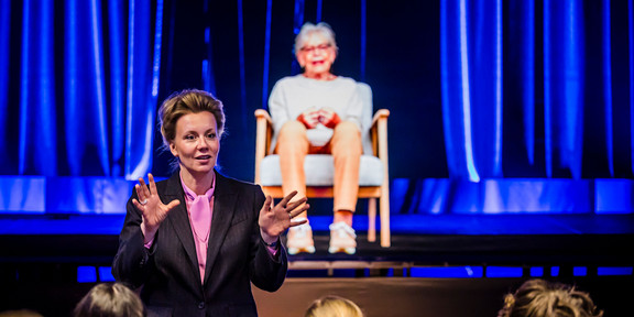 Foto: Eine Person spricht vor einem Publikum, während ein Hologramm auf der Bühne im blauen Licht erscheint.