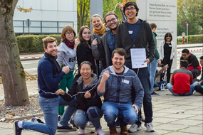 Eine Gruppe internationaler Studierender vor dem Internationalen Begegnungszentrum der TU Dortmund, die ein Spiel spielt; im Hintergrund eine weitere Gruppe