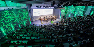 The picture shows the Audimax of TU Dortmund University at the Academic Year Celebration 2024. The photo was taken from above and shows the audience from behind. In the center, further up in the picture, the TU Dortmund University orchestra can be seen, above it the presentation. The side walls of the Audimax are illuminated in green and are also illuminated on both sides with two circles in which the TU logo and the number 24 are displayed.
