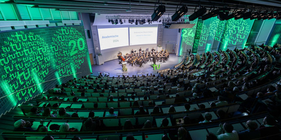 The picture shows the Audimax of TU Dortmund University at the Academic Year Celebration 2024. The photo was taken from above and shows the audience from behind. In the center, further up in the picture, the TU Dortmund University orchestra can be seen, above it the presentation. The side walls of the Audimax are illuminated in green and are also illuminated on both sides with two circles in which the TU logo and the number 24 are displayed.