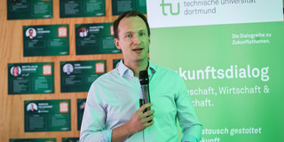Foto: Ein Redner spricht ins Mikrofon, im Hintergrund steht ein Aufsteller des Zukunftsdialogs.