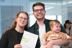 Zwei Personen, die in die Kamera lächeln. Der Mann trägt ein Baby und die Frau hält eine Urkunde in der Hand.
