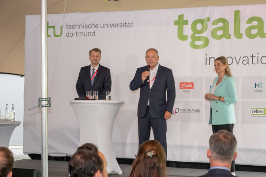 Begrüßung Jan Opländer und Mike Gralla Drei Personen auf einer Bühne mit weißem Hintergrund. Ein Mann hält eine Rede. Auf dem weißen Hintergrund sind diverse Logos zu sehen.