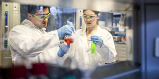 Ein junger Mann und eine junge Frau untersuchen in einem Labor Reagenzgläser mit grüner und roter Flüssigkeit. Sie tragen Schutzkleidung.
