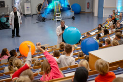 Ein Mann hält in einem vollbesetzten Hörsaal einen großen hellblauen Wasserball in der Hand und übergibt an im Publikum sitzende Kinder.