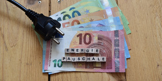 auf Holzdielen liegen Geld und ein Kabel mit Stecker, darauf der Schriftzug "Energiepauschale"