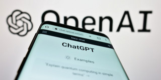 Ein Smartphone, auf dem die Webseite "ChatGPT" geöffnet ist, vor einem Logo von OpenAI.