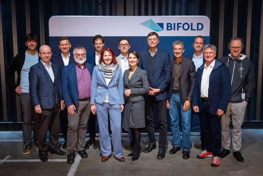 A group of people pose together in front of a canvas stand-up for a photo. On the stand-up is written "BIFOLD".