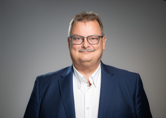 Porträtfoto von Prof. Gerhard Schembecker: Prof. Schembecker trägt einen Anzug und eine blaue Brille. Er steht vor einem grauen Hintergrund und lächelt in die Kamera.
