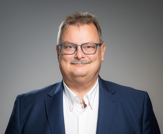 Porträtfoto von Prof. Gerhard Schembecker: Prof. Schembecker trägt einen Anzug und eine blaue Brille. Er steht vor einem grauen Hintergrund und lächelt in die Kamera.