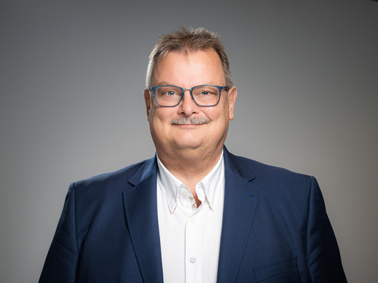 Portrait photo of Prof. Gerhard Schembecker: Prof. Schembecker is wearing a suit and blue glasses. He stands in front of a gray background and smiles at the camera.