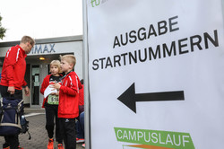 Ein Schild mit der Aufschrift "Ausgabe Startnummer" zeigt einen Pfeil nach links. Im Hintergrund stehen zwei Kinder und ein Mann.