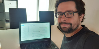 A young man with a beard and glasses sits at a table with his laptop open.