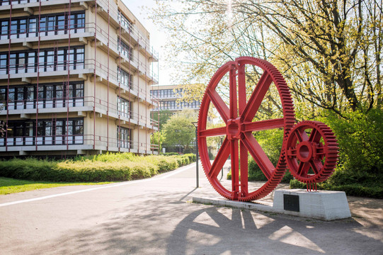 A sculpture of a very large and a smaller red gear stands in front of a multi-story building.