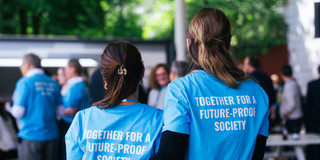 Foto: Zwei Frauen stehen mit dem Rücken zur Kamera und haben die Arme umeinander gelegt. Auf dem Rücken ihrer blauen T-Shirts steht "Together for a future-proof society".