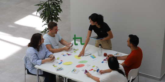 Workshop ZSB 1 Vier Studierende sitzen an einem Tisch. Auf dem Tisch liegen Workshop Materialien, welche von einer Studienberaterin erklärt werden.