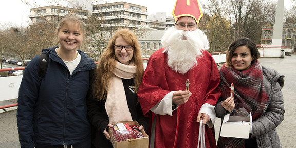 Prof. Ballhorn ist als Nikolaus verkleidet. Drei Studentinnen stehen neben ihm auf der Mensabrücke. Zwei Studentinnen halten kleine Kisten mit Süßigkeiten in der Hand. 
