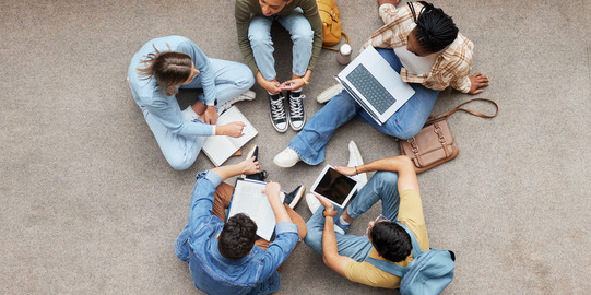 Das Foto zeigt eine Gruppe junger Menschen, die im Kreis auf dem Boden sitzen und zusammen arbeiten oder lernen aus der Vogelperspektive.