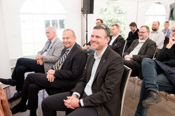 In a tent, several rows of men sit on chairs.