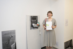 Rundgang Kunst Kunstpreis Plastik Eine Studentin mit Urkunde steht vor einer Installation aus mehreren Grafiken und Fliesenplatten.