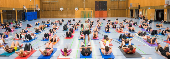 Viele Menschen liegen auf Yogamatten in einer Sporthalle und machen Bauchmuskelübungen unter Anleitung einer Trainerin