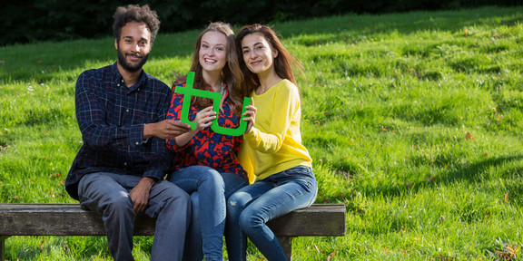 Das Bild zeigt drei junge Erwachsene Personen, davon eine männlich und zwei weiblich gelesene Menschen, wie sie eng zusammen auf einer Holzbank sitzen, gemeinsam das TU-Logo halten und in die Kamera lächeln. Im Hintergrund ist grüne Wiese zu sehen.