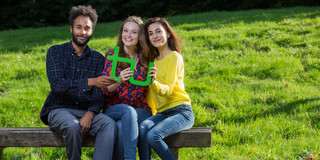 Das Bild zeigt drei junge Erwachsene Personen, davon eine männlich und zwei weiblich gelesene Menschen, wie sie eng zusammen auf einer Holzbank sitzen, gemeinsam das TU-Logo halten und in die Kamera lächeln. Im Hintergrund ist grüne Wiese zu sehen.